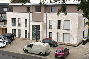 Mehrstöckiges Wohnhaus mit hellbrauner Front und großen Fenstern sowie Parkplatz vor der Tür